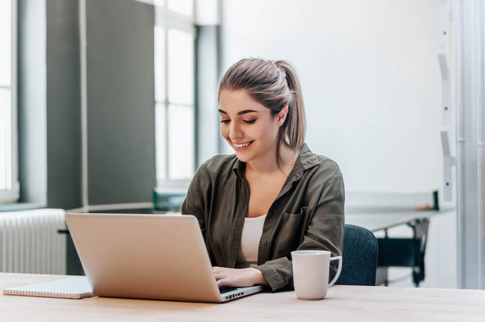 Menina sentada em uma mesa, mexendo em um notebook, sorrindo e com uma canela ao lado.