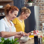 Uma mãe e uma filha, juntas em uma cozinha, preparando alguma refeição enquanto sorriem.
