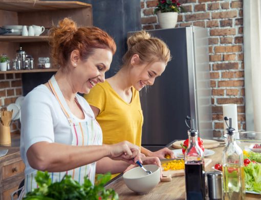 Uma mãe e uma filha, juntas em uma cozinha, preparando alguma refeição enquanto sorriem.