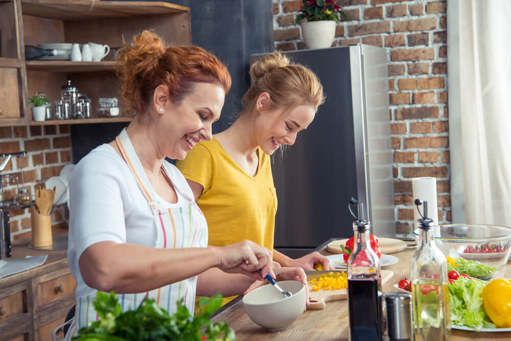 Uma mãe e uma filha, juntas em uma cozinha, preparando alguma refeição enquanto sorriem.